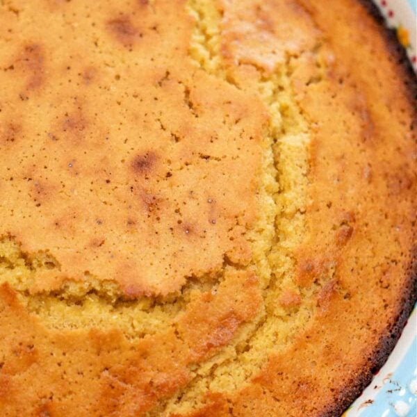 Golden-brown cornbread, a classic Southern dish often made with buttermilk and cornmeal, is shown with a cracked top baked in a round white dish with a scalloped edge.