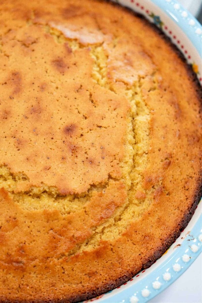 Golden-brown cornbread, a classic Southern dish often made with buttermilk and cornmeal, is shown with a cracked top baked in a round white dish with a scalloped edge.
