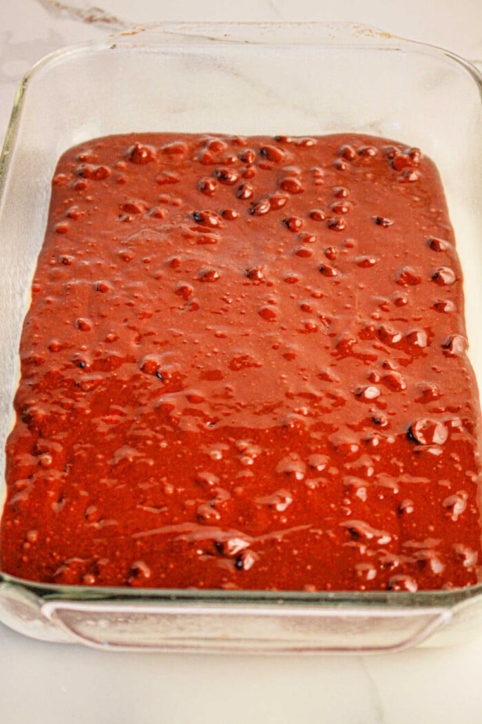 A clear glass baking dish filled with thick, dark chocolate batter looks tempting; it resembles the first step of crafting brookies by combining brownie mix and cookie dough. The batter appears smooth with small, uneven bubbles, ready for baking on the light-colored countertop.