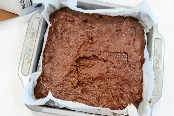 A square baking dish with parchment paper is filled with unbaked pumpkin brownie batter. The batter is spread unevenly across the surface, ready for baking. The pan is labeled "Farberware.