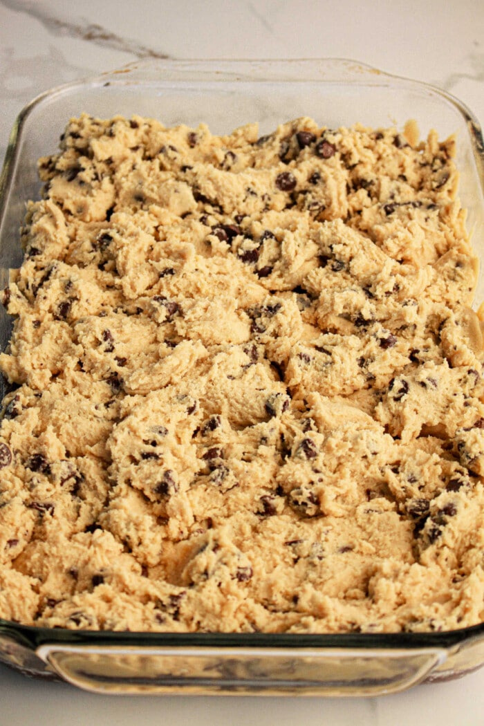 Close-up of a glass baking dish filled with unbaked chocolate chip cookie dough, spread evenly like an easy step in learning how to make brookies with brownie mix and cookie dough. The dough is light brown with visible chocolate chips, ready for baking.