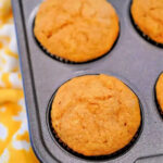 A muffin tin holds six freshly baked golden brown Cake Mix Pumpkin Banana Muffins on a yellow patterned cloth.
