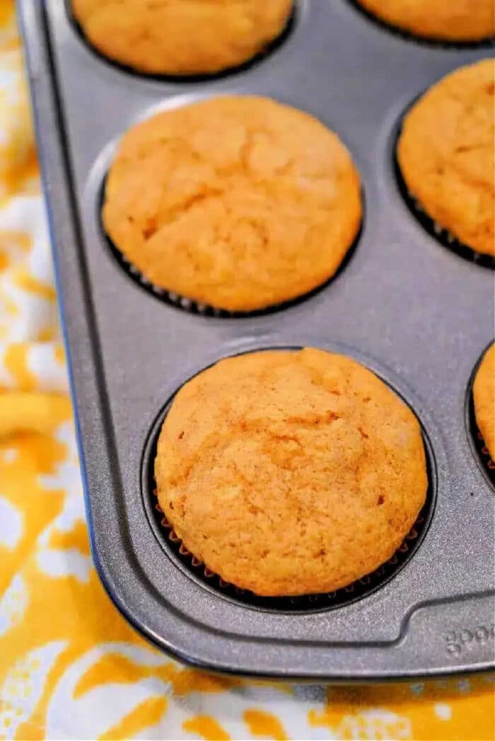 A muffin tin holds six freshly baked golden brown Cake Mix Pumpkin Banana Muffins on a yellow patterned cloth.