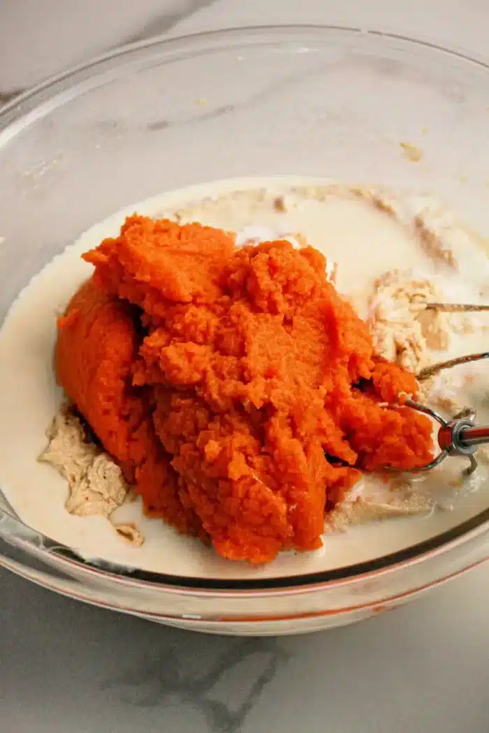 A glass mixing bowl containing a vibrant orange pumpkin puree, pale beige dry ingredients, and a liquid. An electric mixer beater partially submerged in the mixture is visible on the right side of the bowl, ready to transform these elements into a delicious Pumpkin Poke Cake.