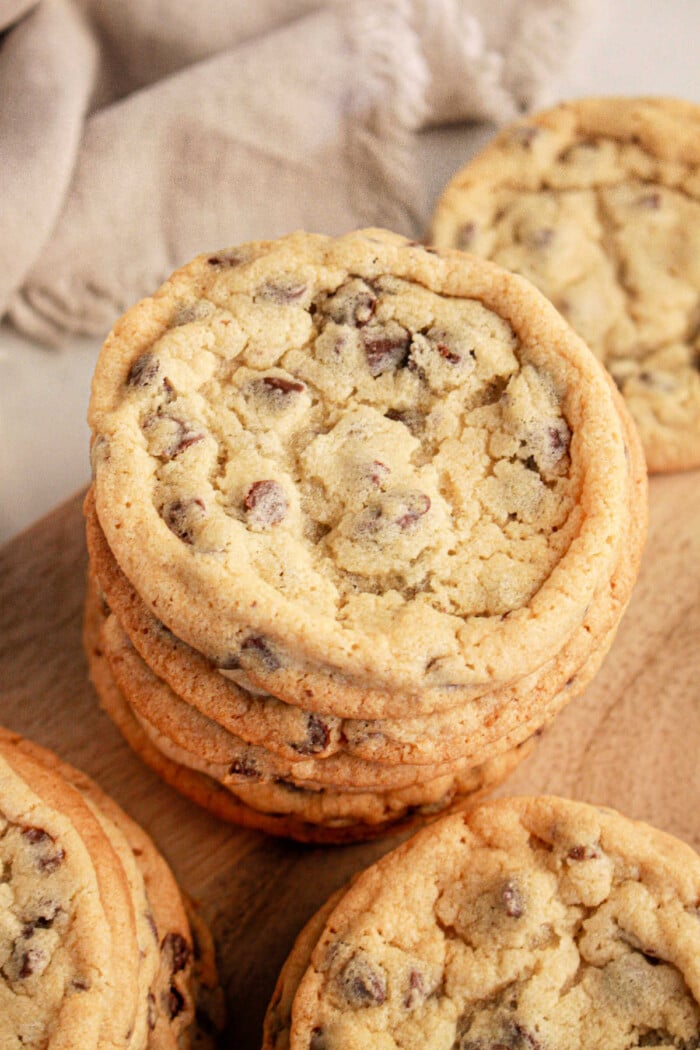 A stack of chocolate chip cookies made with pancake mix sits on a wooden surface. The cookies boast a golden brown color with visible chocolate chips and a slightly cracked texture on top. A beige cloth adds a cozy touch in the background.