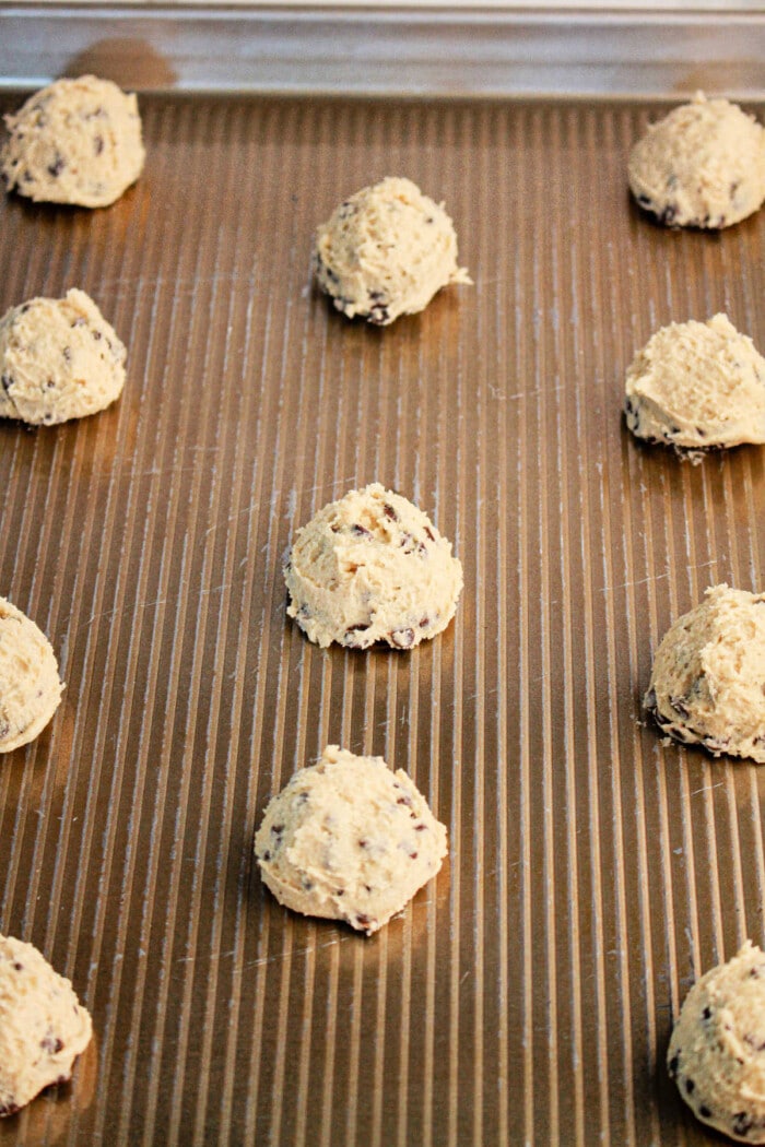 Scoops of cookie dough, crafted from pancake mix, are evenly spaced on a ribbed baking sheet, ready for baking. The dough contains visible chocolate chips.