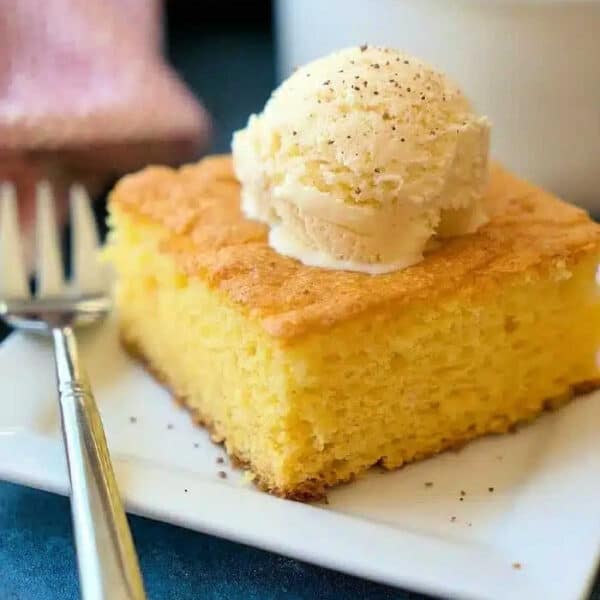 A slice of egg nog pudding cake topped with a scoop of vanilla ice cream sits on a square white plate with a fork.