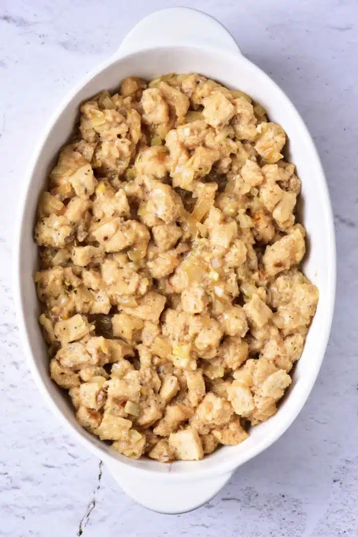 A white oval dish filled with bread stuffing, containing pieces of bread mixed with herbs and onions, rests on a light-colored marble surface, illustrating how to cook stuffing in the oven.