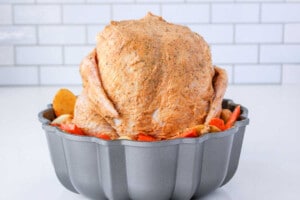 A whole seasoned chicken sits upright in a bundt pan, surrounded by perfectly sliced potatoes and carrots. Wondering how long to cook a whole chicken like this? The backdrop is a simple white tile wall, adding an elegant touch.