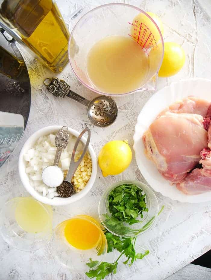 A delightful setup for easy chicken recipes awaits, with raw chicken, lemon juice, olive oil, and chicken broth elegantly placed on a white textured surface. Chopped onions, garlic, cilantro, measuring spoons are ready to assist while vibrant lemons and parsley add a fresh touch nearby.