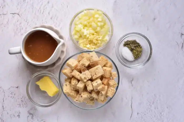 Ingredients for how to cook stuffing in the oven are laid out on a white textured surface: a pitcher of brown liquid, diced onions and garlic in a small bowl, dried herbs with salt in another, croutons in a medium bowl, and a pat of butter resting on a small plate.