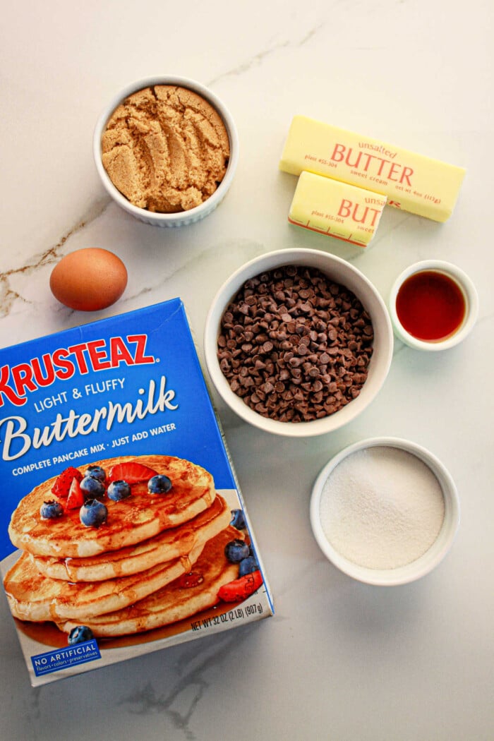 Ingredients for a baking recipe are laid out on a counter: a box of pancake mix perfect for cookies, a bowl of brown sugar, chocolate chips, two sticks of butter, vanilla extract in a small cup, white sugar in another bowl, and one egg.