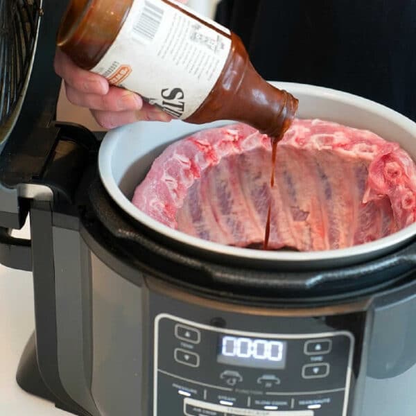 Person pouring barbecue sauce onto raw ribs in a Ninja Foodi, getting the Ninja Foodi ribs ready to be cooked to tender perfection.