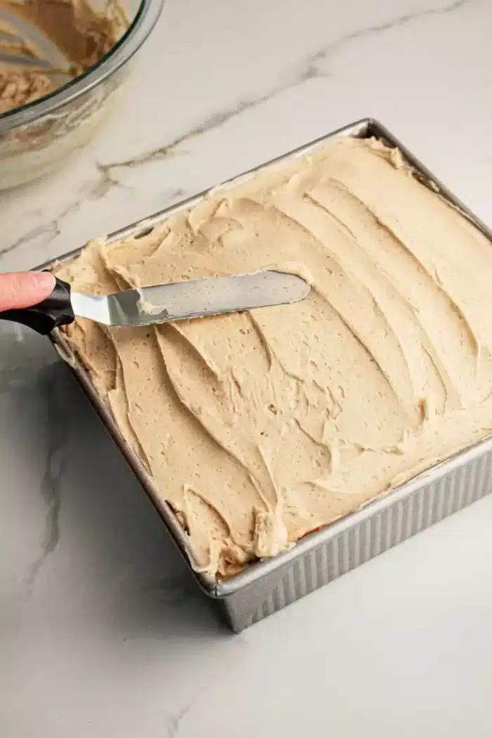 A hand holding a straight icing spatula is spreading beige frosting evenly over a square Pumpkin Poke Cake in a metal baking pan. The pan is sitting on a marble countertop, and a glass bowl with additional frosting is visible in the background.