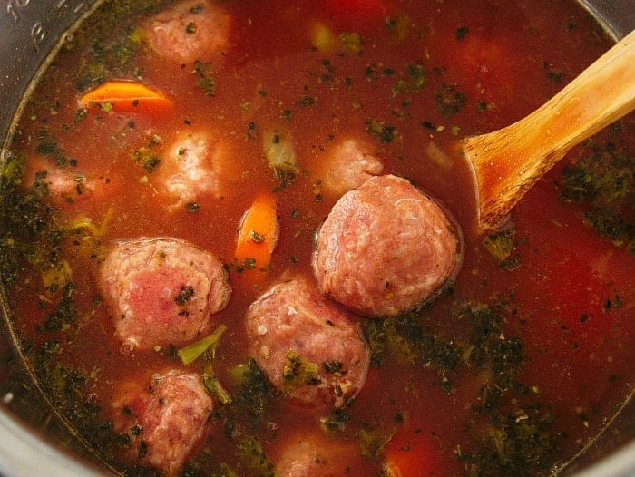 Instant Pot meatball soup features meatballs, carrots, and herbs simmering in a tomato-based broth, stirred with a wooden spoon.