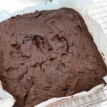 A freshly baked square chocolate cake, infused with hints of pumpkin to resemble rich brownies, sits in a baking tray lined with parchment paper, cooling on a wire rack.