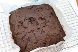 A freshly baked square chocolate cake, infused with hints of pumpkin to resemble rich brownies, sits in a baking tray lined with parchment paper, cooling on a wire rack.