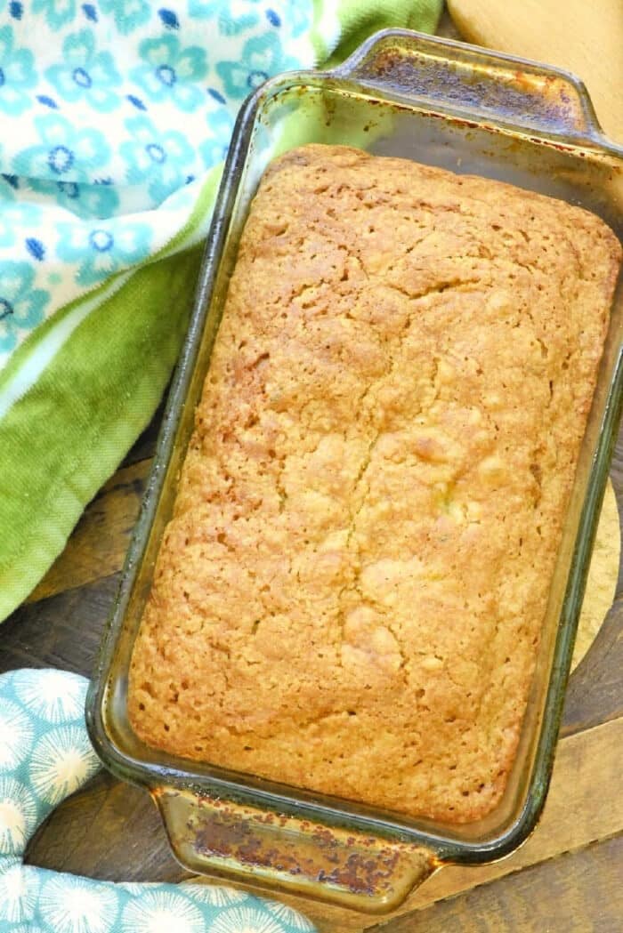 Zucchini pineapple bread is so amazing!! The best way to make zucchini bread super moist and brings in a great sweetness naturally with added pineapple. A loaf of baked zucchini pineapple bread in a glass pan rests on a wooden surface with patterned towels nearby.