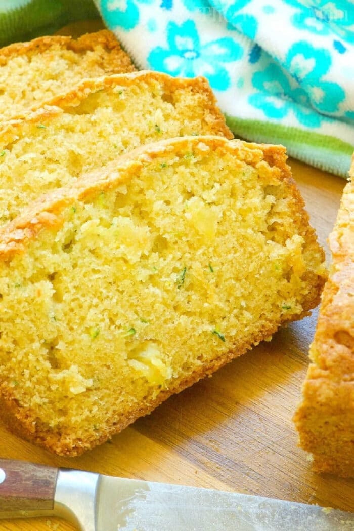 Zucchini pineapple bread is so amazing!! The best way to make zucchini bread super moist and brings in a great sweetness naturally with added pineapple. Close-up of sliced zucchini pineapple bread on a wooden surface, with a blue floral towel in the background.