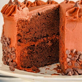 Brownie cake with chocolate frosting and decorative chocolate chips on a white cake stand. A slice is removed, showcasing the rich texture of the brownie mix.