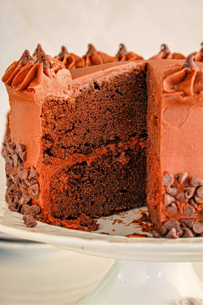 Brownie cake with chocolate frosting and decorative chocolate chips on a white cake stand. A slice is removed, showcasing the rich texture of the brownie mix.