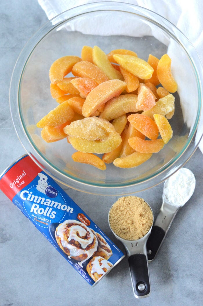 A bowl of frozen sliced peaches, cinnamon rolls can, brown sugar, and flour sit on a gray surface&mdash;perfect for making an easy Cinnamon Roll Peach Cobbler.