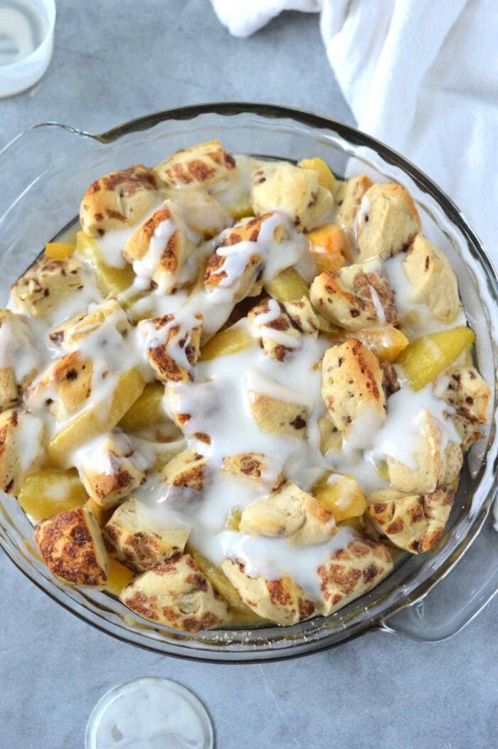 A glass dish filled with Cinnamon Roll Peach Cobbler, featuring cinnamon roll pieces, peach slices, and white icing on top.