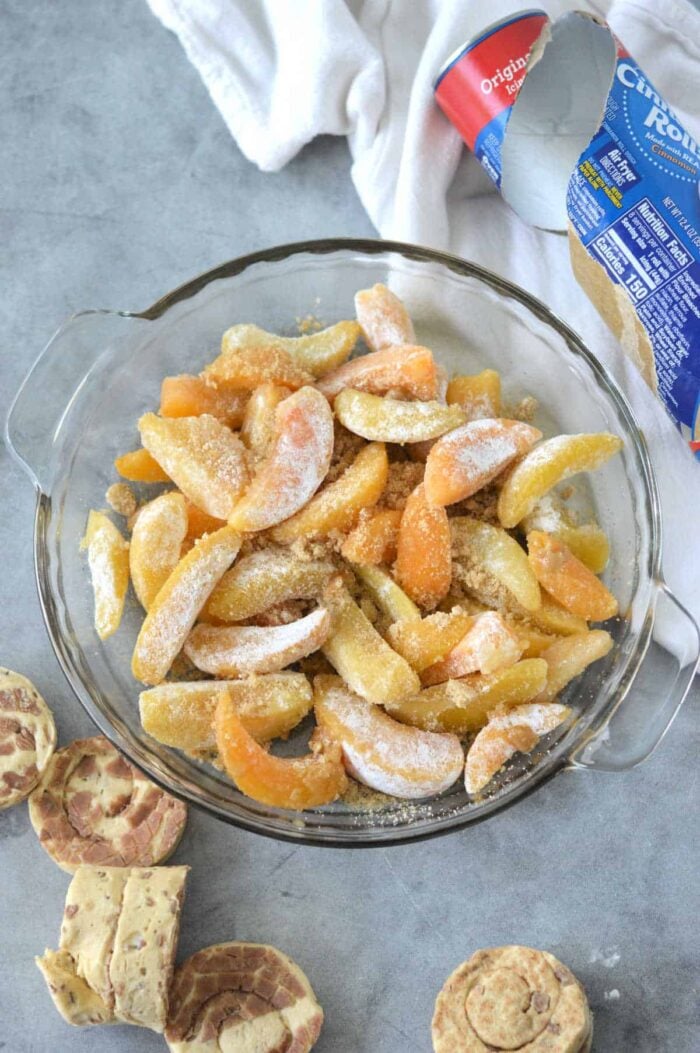 Sliced frozen peaches and apples with sugar in a pie dish, perfect for making Cinnamon Roll Peach Cobbler, with biscuit dough and wrapper nearby on a gray surface.