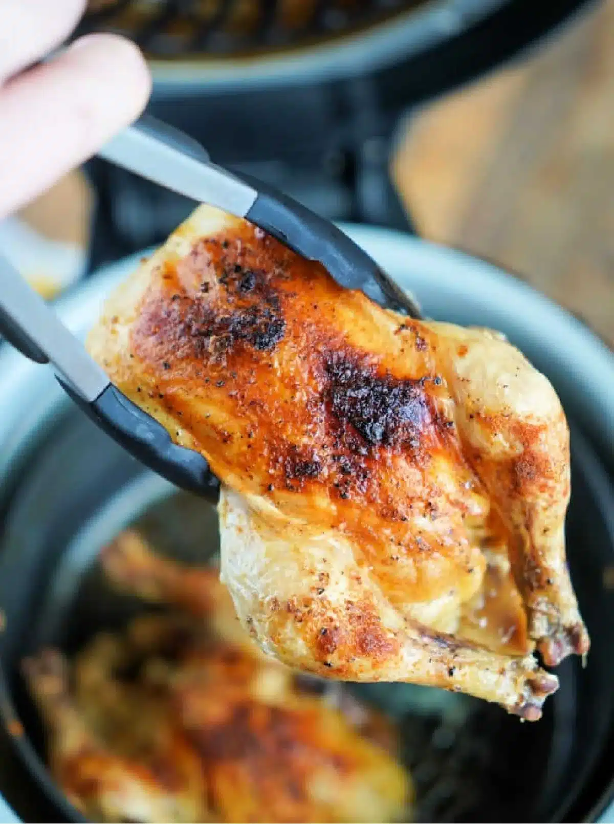 A cooked Cornish hen is being held with tongs above an air fryer. The skin is golden brown and slightly crispy. In the background, another hen roasts to perfection inside the air fryer, showcasing one of many delightful Ninja Foodi recipes.