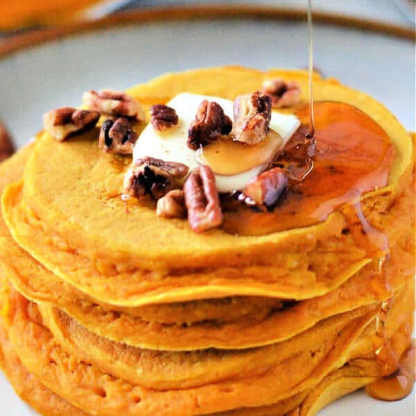 A stack of Pumpkin Pancakes with Bisquick topped with butter, chopped pecans, and syrup being poured over them.