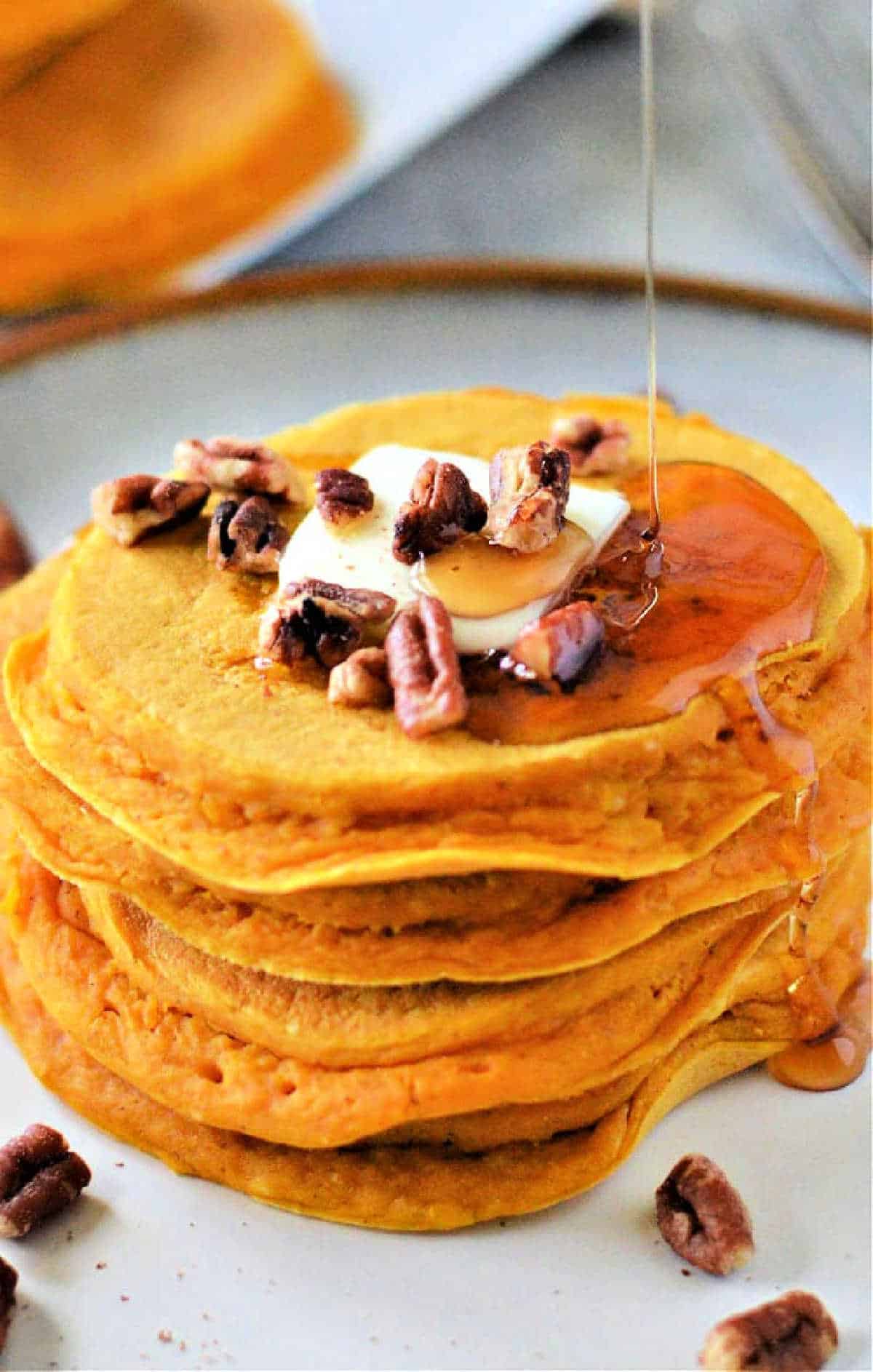 A stack of Pumpkin Pancakes with Bisquick topped with butter, chopped pecans, and syrup being poured over them.