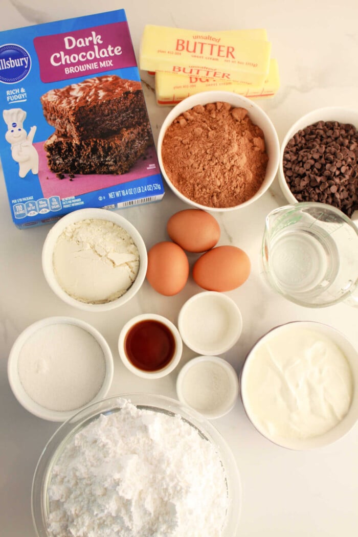 Ingredients for a rich brownie cake: eggs, butter, chocolate chips, flour, sugar, vanilla, yogurt, and water are all displayed on the table.