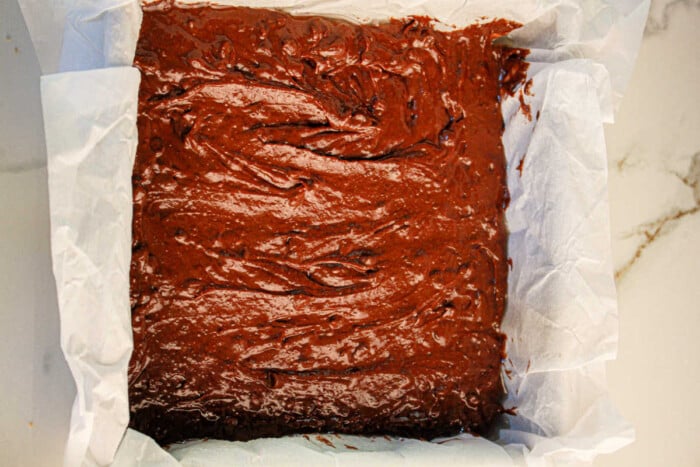 A parchment-lined square baking tin rests on a marble surface, brimming with rich chocolate black bean brownie batter, ready to mix magic in the oven.