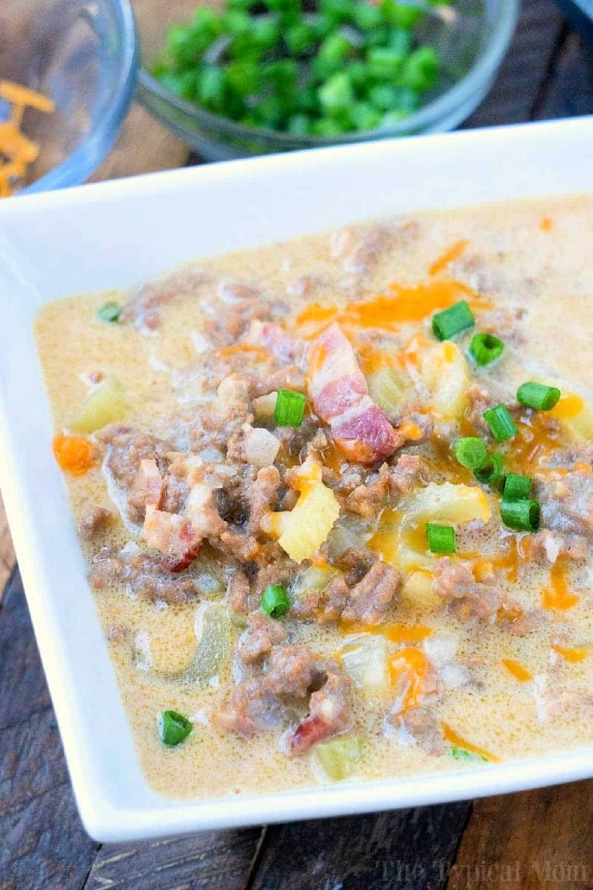 Instant Pot cheeseburger soup is so good! It's a fun pressure cooker meal that your kids will love and will make you come back for seconds. #instantpot #cheeseburger #soup #pressurecooker #groundbeef #instantpotrecipes #instantpotsoup #thetypicalmom Creamy instant pot cheeseburger soup in a white bowl, topped with ground beef, crispy bacon, melty cheese, and chopped green onions.