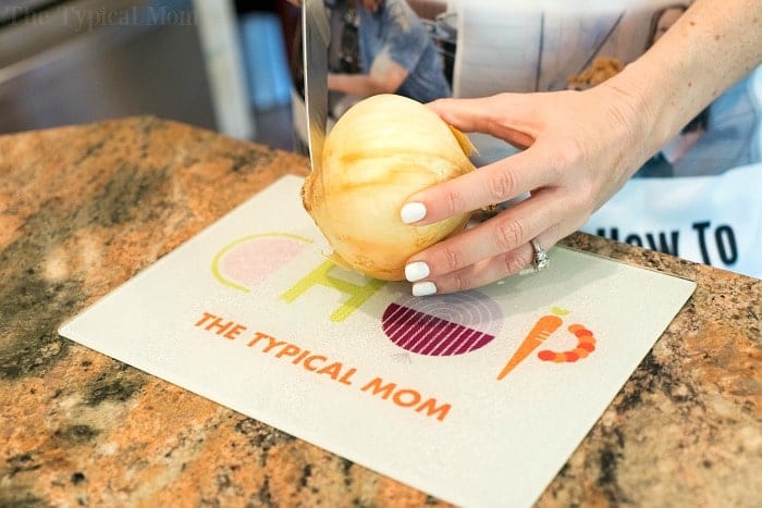 A person with white nail polish is slicing an onion on a cutting board marked with "CHOP THE TYPICAL MOM," preparing ingredients for an instant pot cheeseburger soup. The cutting board rests elegantly on a granite countertop.