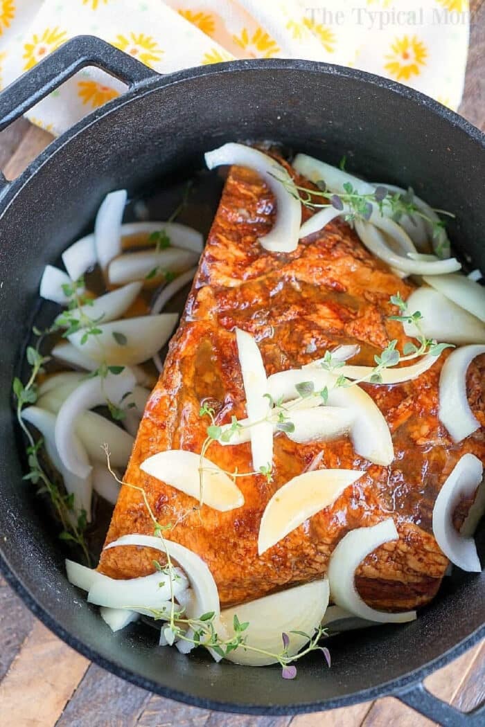 This dutch oven ham needs to go on your list of the best dutch oven recipes this year, and with a honey glaze it is perfect for Christmas or Easter dinner. #dutchoven #ham #dutchovenrecipes #honeyham Seasoned dutch oven ham in a black pot, topped with sliced onions and fresh herbs, ready to be cooked.