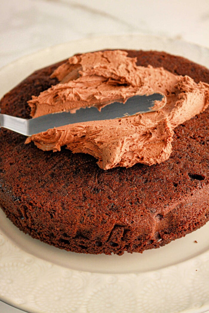 A knife gracefully spreads chocolate frosting on a round brownie cake, crafted from rich brownie mix, resting elegantly on a white plate.