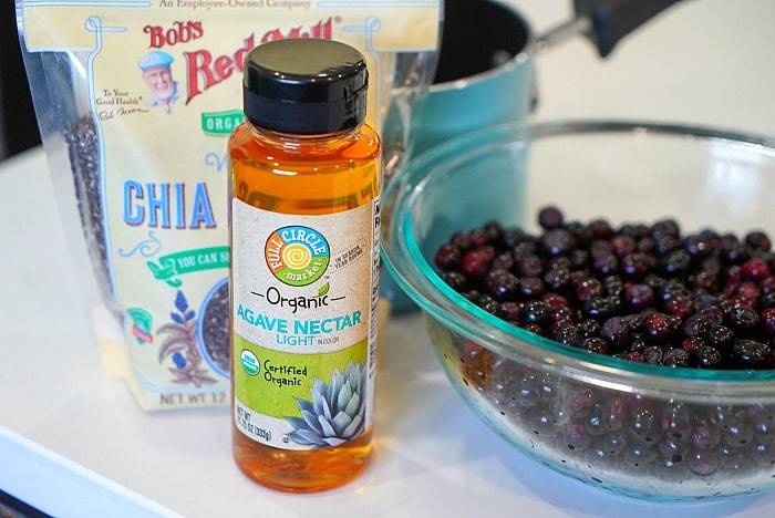 A bottle of agave nectar, a bag of chia seeds, a bowl of fresh berries, and a jar of huckleberry jam sit on the kitchen counter.
