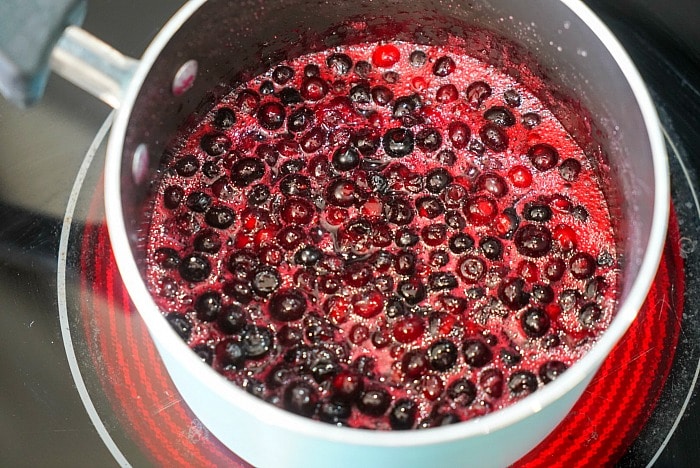 Blueberries cooking in a saucepan, forming a bubbling fruit sauce on a stovetop reminiscent of homemade huckleberry jam.