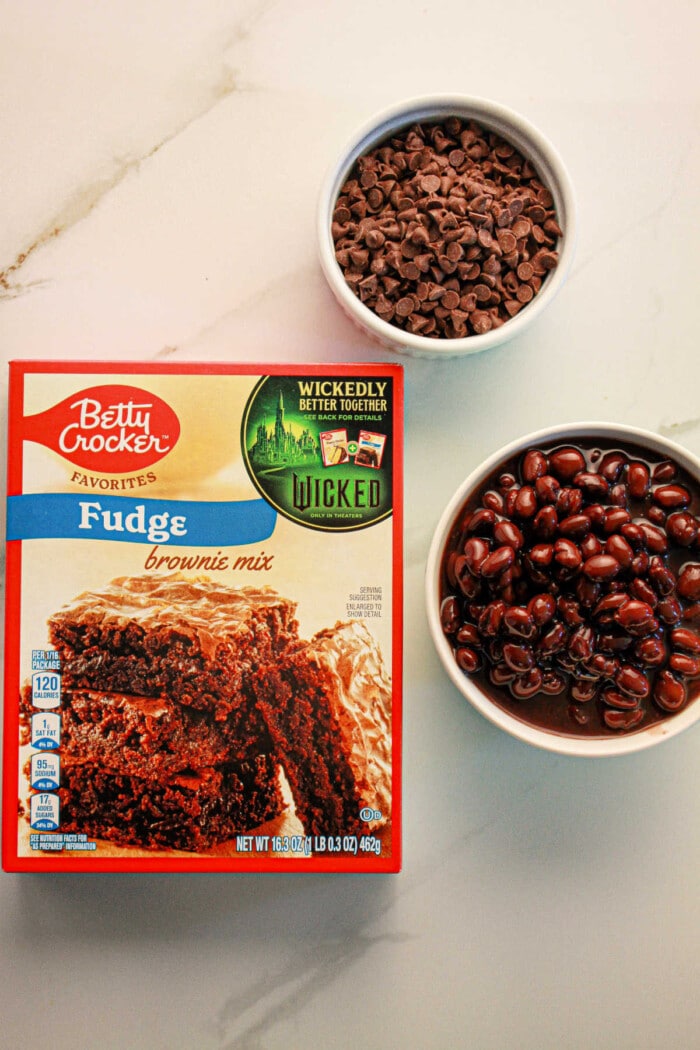 A box of fudge brownie mix sits alongside bowls of chocolate chips and black beans, perfectly set up to create delicious black bean brownies with mix on a pristine white surface.