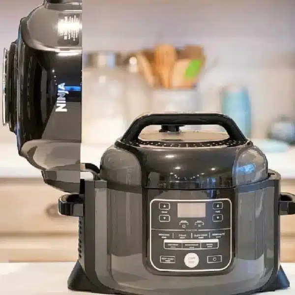 A black multicooker with an open lid sits on a kitchen counter, ready for your favorite Ninja Foodi recipes. The appliance features control buttons and a digital display on its front. In the background, blurred kitchen items like jars and utensils add a cozy touch to the scene.