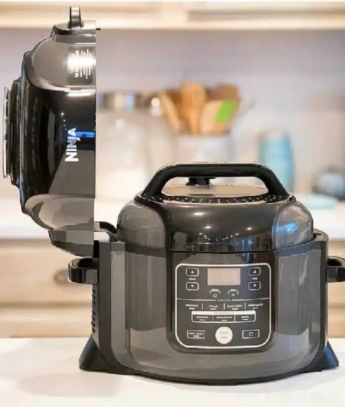 A black multifunctional kitchen appliance, perfect for crafting Ninja Foodi recipes, sits on the counter. The lid is open, revealing the inner cooking chamber. The control panel with digital display and various buttons is visible, while blurred kitchen utensils and jars linger in the background.