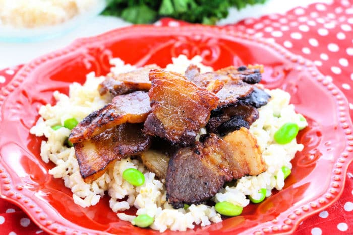 Crispy bacon pieces&mdash;perfect for those learning how to cook pork belly in the oven&mdash;are served on a bed of rice with green edamame on a red plate.