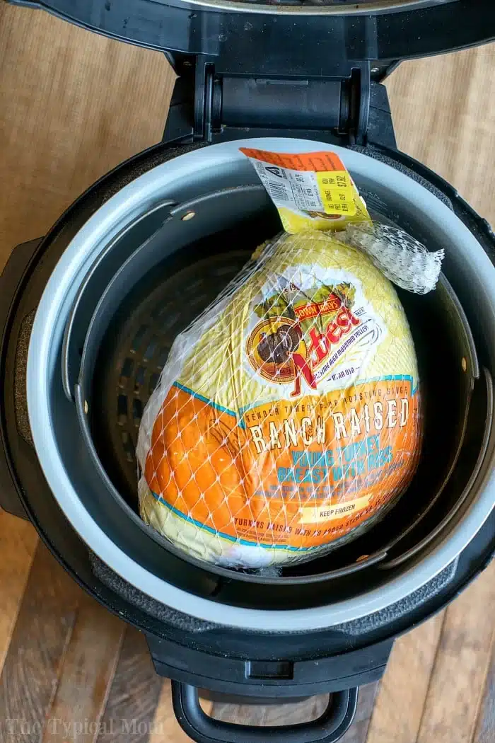 Ninja foodi turkey breast that's tender inside and crispy on the outside! Learn how to cook a bone in turkey breast in your Ninja Foodi or Instant Pot. #instantpotrecipes #ninjafoodirecipes #pressurecooker #turkeybreast #turkey #thetypicalmom #ninjafoodi #instantpot A frozen whole turkey in packaging sits inside an open Instant Pot pressure cooker, ready to transform into a juicy pressure cooker turkey breast on a wooden surface.