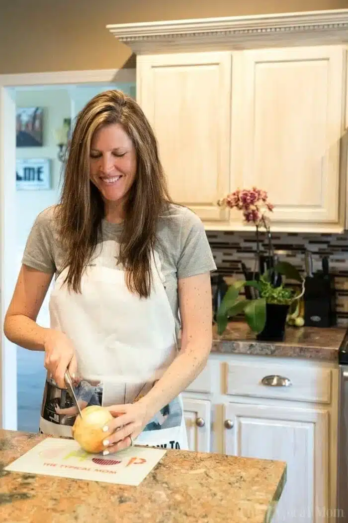 A woman with long hair stands in the kitchen, smiling as she slices a large onion on a cutting board. Dressed in a gray t-shirt and white apron, she's preparing ingredients for an instant pot cheeseburger soup. Plants and kitchen items line the countertop behind her.