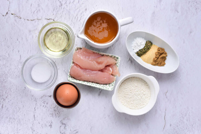 Raw chicken, an egg, flour, seasonings, oil, salt, and broth in bowls on a light textured surface&mdash;perfect for making crispy beer battered chicken tenders.