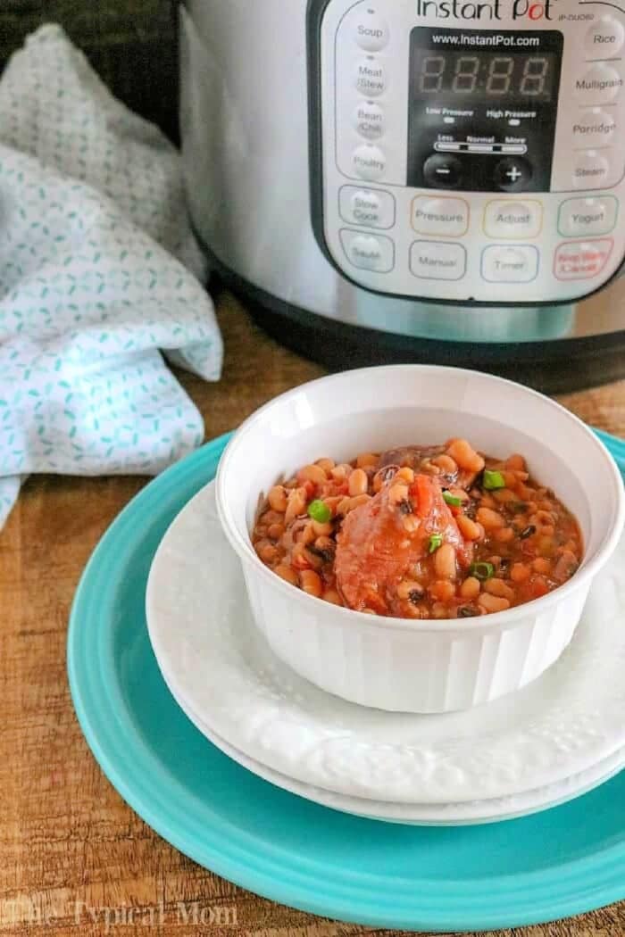 Here is an easy Instant Pot black eyed peas and ham recipe as well as how to make it in a crockpot too. How to make slow cooker black eyed peas and ham using dry beans is great to have on hand after the holidays when you have a lot of leftover ham! Makes for a hearty and healthy meal that everyone will love. #instantpot #pressurecooker #crockpot #slowcooker A white bowl of bean stew sits on plates in front of an Instant Pot, featuring hearty pressure cooker black eyed peas and ham, on a wooden surface.
