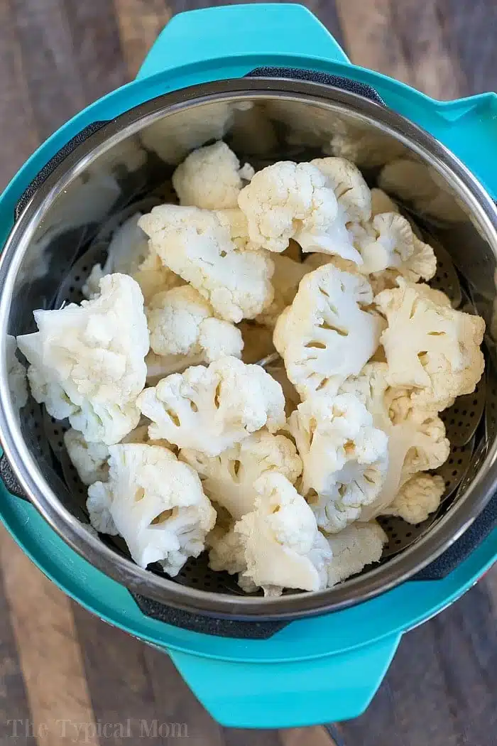This cheesy Instant Pot cauliflower is a great low carb macaroni and cheese style side dish everyone in your family will love, and want you to make again. #instantpot #pressurecooker #lowcarb #sidedish #cauliflower #macaroniandcheese #instantpotrecipes #thetypicalmom Cheesy Instant Pot cauliflower florets in a metal steamer basket inside a blue pressure cooker, ready for cooking.