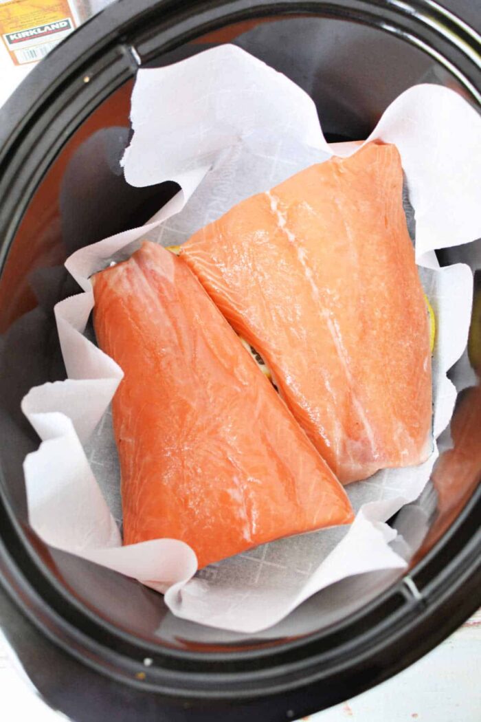 Two raw Crockpot Salmon fillets rest on parchment paper inside a slow cooker, ready to be cooked.