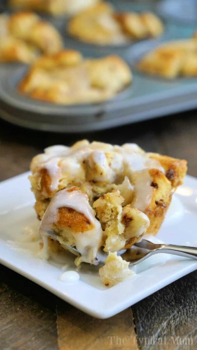 A cinnamon roll muffin with icing on a white plate and a fork, surrounded by apple pie monkey bread muffins in the background.